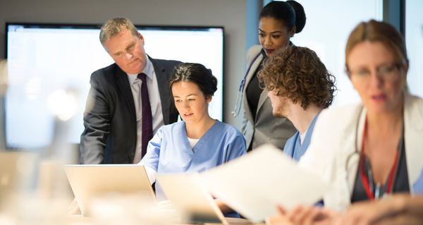 Klinikteam in blauer Dienstkleidung bespricht gemeinsam Unterlagen an einem Laptop