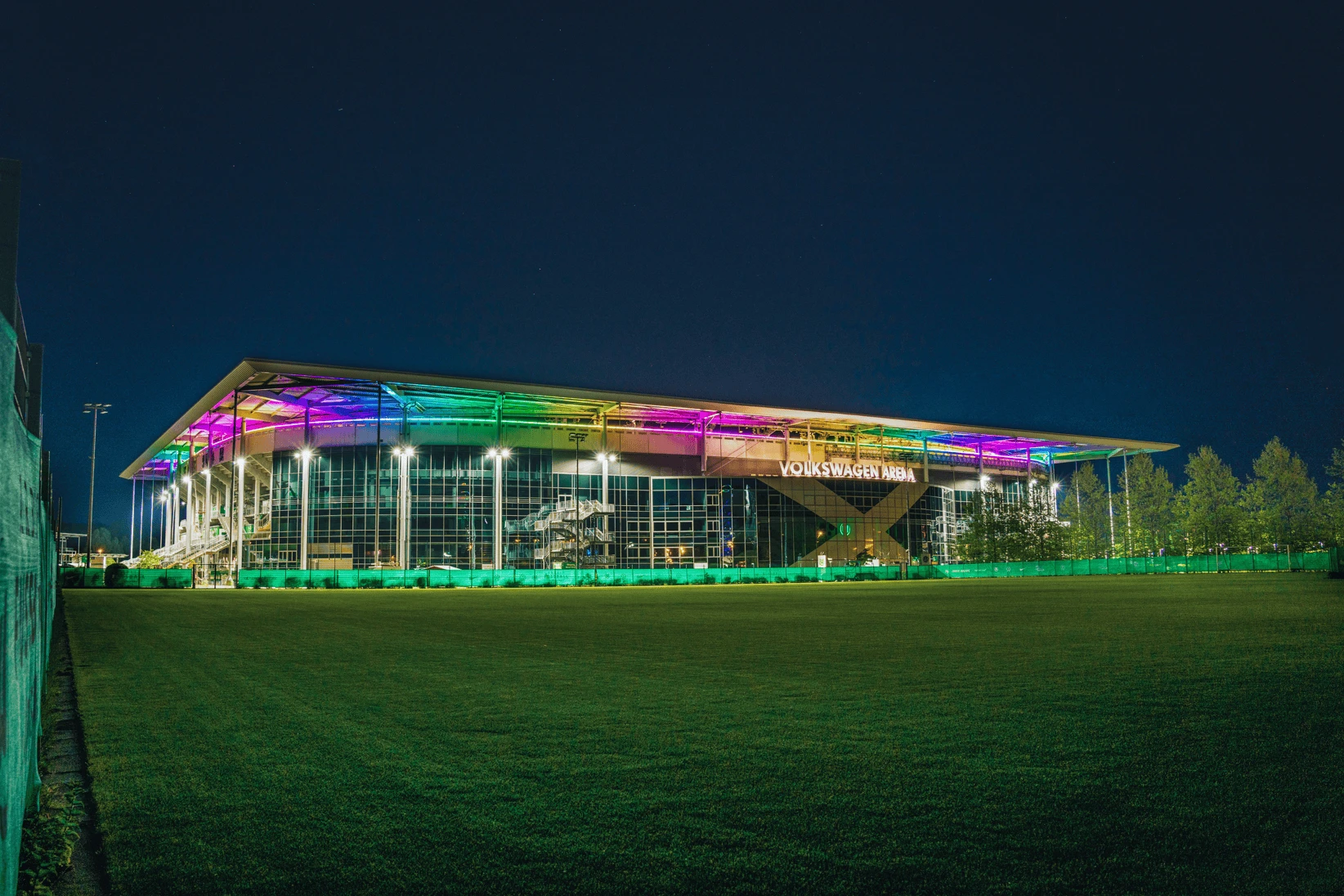 Volkswagen Arena bei Nacht mit farbig beleuchtetem Dachrand hinter einem Rasenplatz