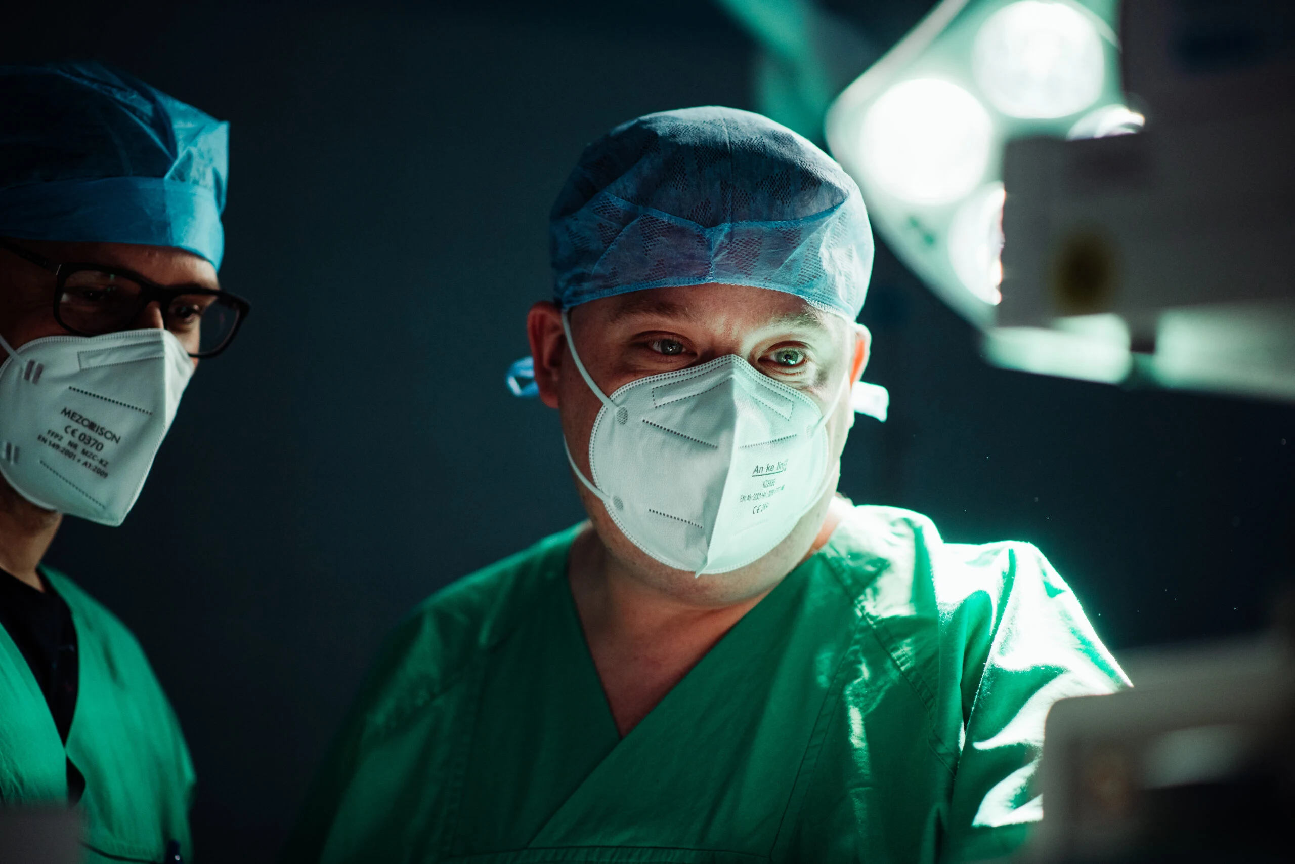 Two people in surgical attire with face masks in the operating room under a surgical light