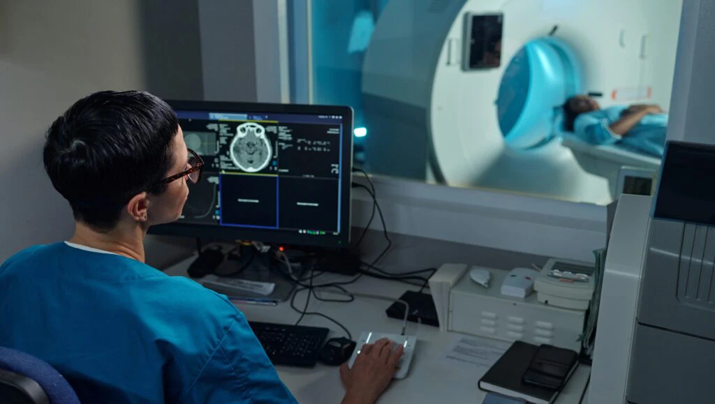 Medizinische Fachkraft am Bildschirm einer CT-Untersuchung neben dem Scanner